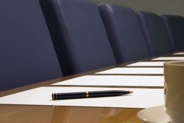 A close-up of a conference room showing a row of chairs, a table, part of a coffee cup, documents (blank) and a pen resting on a piece of paper. Sharp focus on the pen.